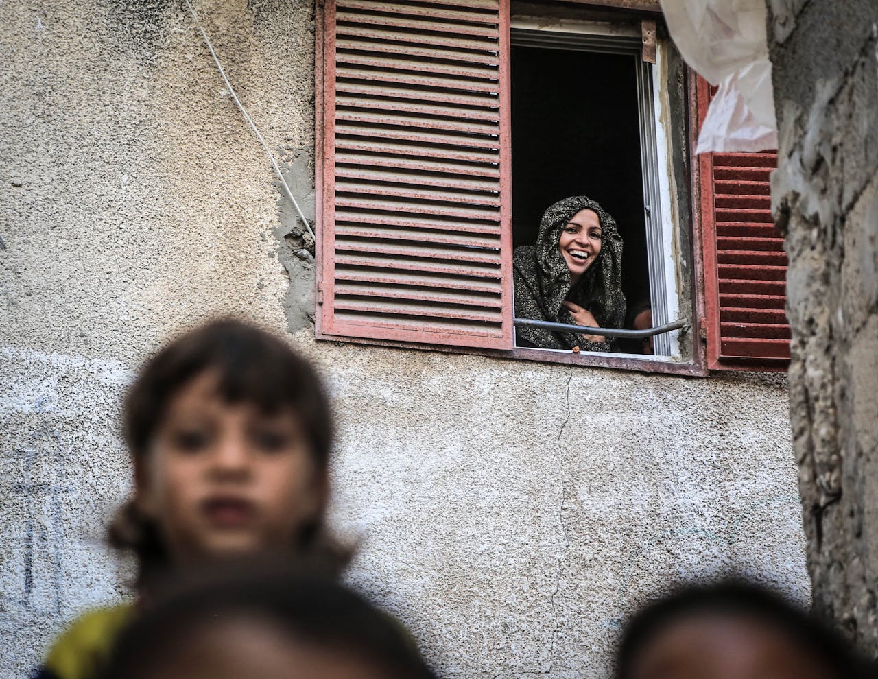 A joyful woman wearing a headscarf smiles from her window above the street.